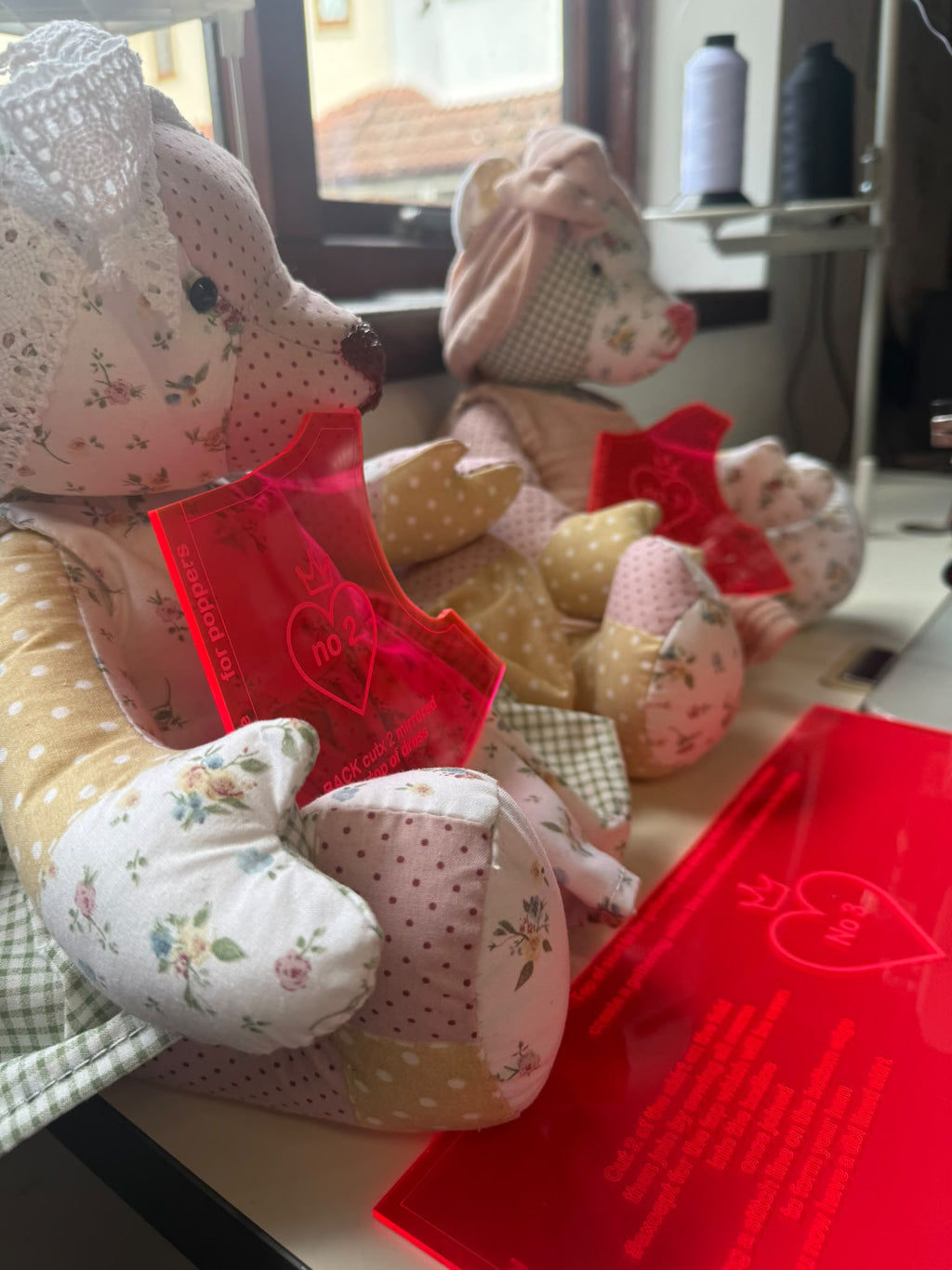 Two patchwork bears wearing floral dresses beside pink acrylic pieces, showing finished dress examples.